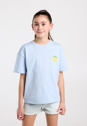 Jong meisje met bruin haar in een paardenstaart, een lichtblauw T-shirt met een citroendessin en lichte spijkershorts, staand tegen een effen achtergrond.