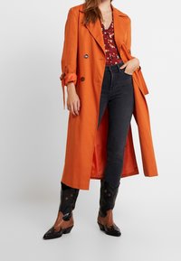 Largo abrigo naranja tipo trench con un diseño a medida, botones negros, combinado con una blusa floral, jeans oscuros y botas de vaquero en negro y marrón.