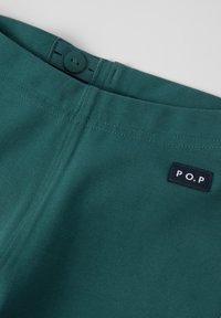 Teal puuvillakangas, jossa on nappikiinnitys vyötäröllä ja musta kangastunnus valkoisilla kirjaimilla "P.O.P." sivussa.