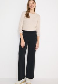 Femme souriante aux longs cheveux bruns, portant un pull beige, un pantalon large noir et des baskets blanches, sur un fond blanc uni.