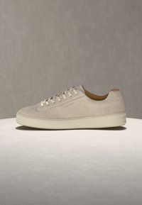 Ανοιχτό μπεζ σουέτ sneaker χαμηλού ύψους με λευκά κορδόνια και χοντρή, επίπεδη κρεμ χρώματος σόλα σε ουδέτερο φόντο.