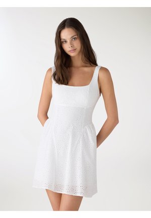 Donna con capelli castani lunghi che indossa un vestito bianco senza maniche con scollo quadrato, in piedi davanti a uno sfondo bianco uniforme.