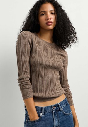 Giovane donna con capelli ricci che indossa un maglione marrone a coste aderente e jeans blu con le mani nelle tasche su uno sfondo semplice.