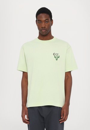 T-shirt in cotone verde chiaro con maniche corte, colletto tondo e grafica di un cactus sul lato sinistro del petto. Vestibilità rilassata, texture morbida.