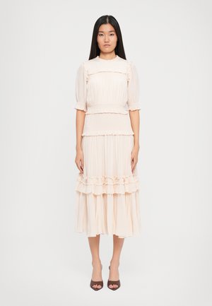 Jonge vrouw staat in een lichtroze midi-jurk met ruffles en pofmouwen, met bruin open teenhakken, tegen een effen witte achtergrond.