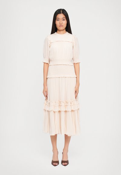 Une jeune femme se tient debout, portant une robe midi à volants de couleur rose pâle avec des manches bouffantes et des sandales à talons ouverts marron, devant un fond blanc uni.