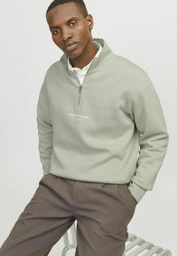 Light green zip-up sweater made of soft fabric with a high collar, featuring a logo in white. Paired with dark brown trousers.