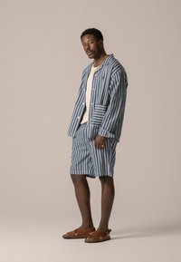 Camicia a maniche corte a strisce blu e bianche e pantaloni corti coordinati, realizzati in tessuto leggero. Sandali marroni completano l'outfit.
