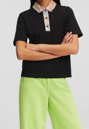 Personne aux bras croisés portant un t-shirt noir à manches courtes avec un col texturé multicolore et un pantalon vert néon.