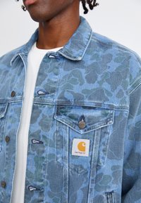 Blaue Jeansjacke mit einem Camouflage-Muster in verschiedenen Blau- und Grüntönen, ausgestattet mit Metallknöpfen und einem Markenpatch auf der Tasche.