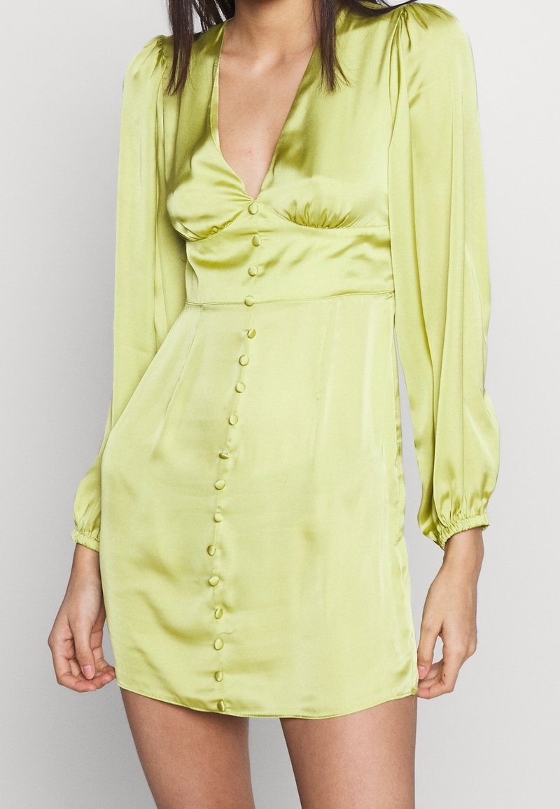 Femme portant une mini-robe en satin vert clair avec de longues manches bouffantes et une rangée de boutons recouverts de tissu à l'avant.