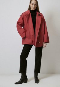 Giacca rossa oversize con spalline, due tasche anteriori e grandi bottoni, abbinata a un dolcevita nero e pantaloni neri a sigaretta.