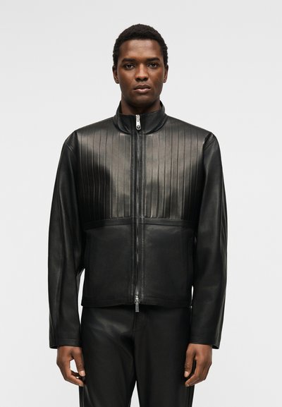 Άνδρας φοράει μαύρο δερμάτινο μπουφάν με φερμουάρ και κάθετες ραφές στο στήθος και μαύρο δερμάτινο παντελόνι, με μονόχρωμο φόντο.