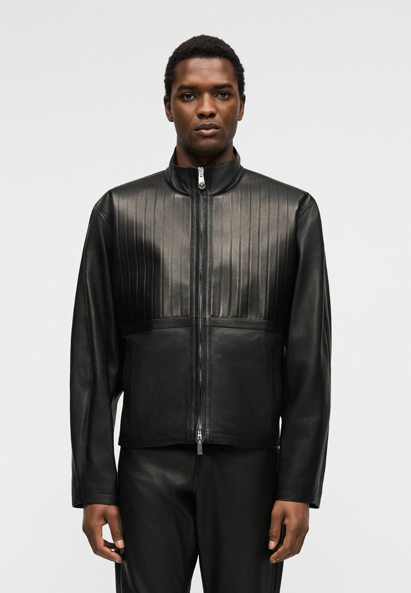 Homme portant une veste en cuir noire zippée avec des coutures verticales sur la poitrine et un pantalon en cuir noir assorti, sur un fond uni.