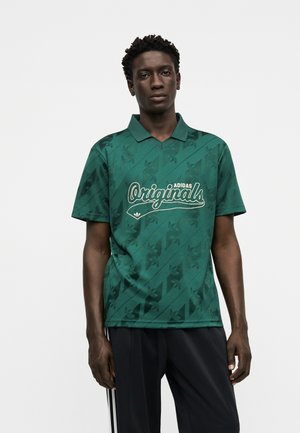 Homme portant une chemise à col Adidas Originals vert foncé avec un motif géométrique et un pantalon noir à bandes blanches, debout devant un fond uni.