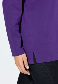 Lange mouwen paarse T-shirt van lichtgewicht stof, met een zijsplit aan de onderkant en een ronde hals. Gewaden over donkere jeans.