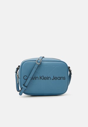 Sac bandoulière rectangulaire bleu avec sangle ajustable, portant l'inscription « Calvin Klein Jeans » sur le devant et un logo « CK » en relief discret.