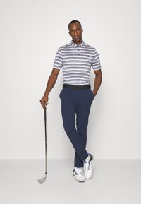 Námořnické modré golfové kalhoty ve spojení s šedobílým pruhovaným polo tričkem, které má límec a tři knoflíky. Model drží golfovou hůl.