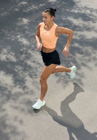 Tenue de sport comprenant un crop top sans manches orange clair, un short noir à rayures et des chaussures de course vert clair, le tout sur un fond de pavé lisse.