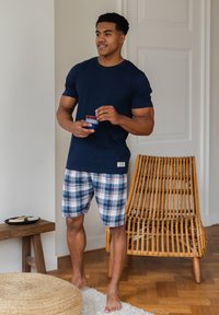 T-paita, jossa lyhyet hihat tummansinisenä, yhdistettynä ruudullisiin shortseihin, joissa on sinistä, valkoista ja punaista. Mies pitää kuppia kädessään seisten puulattialla.