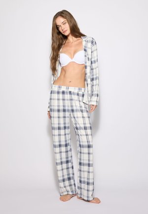 Giovane donna in piedi scalza, indossa un reggiseno bianco e un completo pigiama a quadri bianco con camicia aperta su uno sfondo chiaro uniforme.