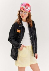 Czarna dżinsowa kurtka z przednimi kieszeniami i pomarańczową naszywką graficzną, biała koszulka pod spodem oraz żółte lniane szorty. Kolorowa kwiatowa chustka na głowę.