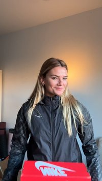 Jachetă neagră ușoară cu mâneci cu puncte, purtată peste un top negru. Cutie de pantofi Nike roșie vizibilă în prim-plan. Iluminare naturală în fundal.