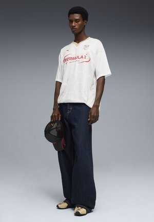 Joven de pie con camiseta blanca de la Formula 1 Racing League, vaqueros anchos oscuros, zapatillas beige, sosteniendo una gorra negra y roja en su mano derecha.