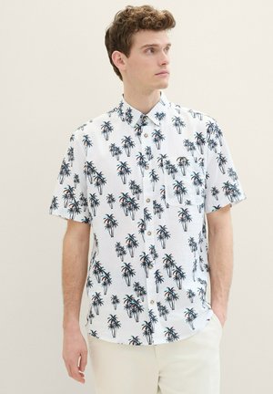 Chemise à manches courtes en tissu blanc avec un imprimé de palmiers en vert et marron. Elle comporte une poche avant et un col classique.