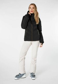 Giacca nera isolata con colletto alto, tasche con zip e design aderente, abbinata a pantaloni chiari e stivali da sci grigi.