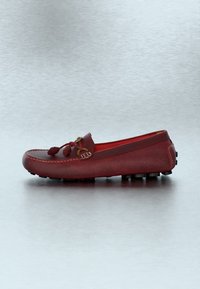 Chaussure mocassin en cuir rouge avec un détail de lacet noué et des semelles en caoutchouc antidérapantes, présentée en profil sur un fond gris clair.
