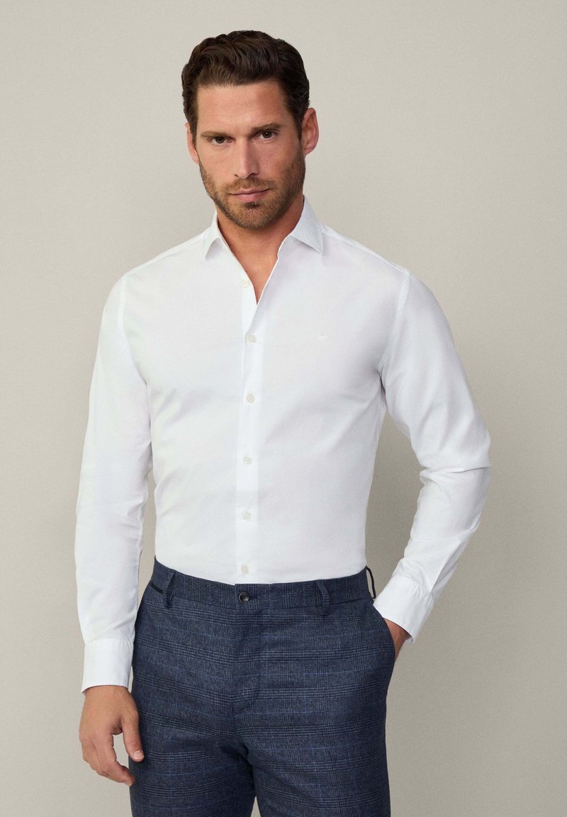 Camicia bianca a maniche lunghe con chiusura a bottone e colletto morbido, abbinata a pantaloni blu testurizzati con un delicato motivo a quadretti.