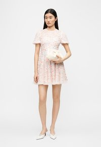 Γυναίκα που φοράει κοντό, παστέλ φλοράλ φόρεμα με φουσκωτά μανίκια, κρατώντας ένα λευκό τσαντάκι και φορώντας λευκά μυτερά τακούνια.