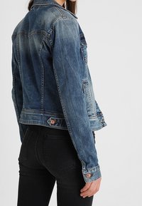 Denim jacket in faded blue with a classic cut, front seams, and button accents at the cuffs and waistband. Textured fabric with a worn look.