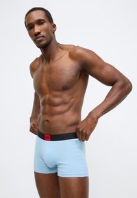 Ανοιχτό μπλε ανδρικά εσώρουχα σόρτς από βαμβακερό μείγμα, με μαύρη ελαστική μέση και κόκκινη ετικέτα λογότυπου. Ο σχεδιασμός είναι εφαρμοστός και λείος.