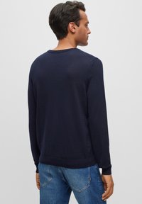 Jūriniu stiliaus apvalios apykaklės megztinis, su megzto audinio tekstūra, ilgos rankovės, su raukiniais ant rankovių ir apatinės dalies, dėvimas su mėlynomis džinsinėmis kelnių, matomas iš nugaros.