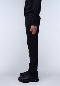 Black cargo pants with a slim fit, featuring multiple pockets and a durable fabric. Paired with black slip-on shoes, side profile view.