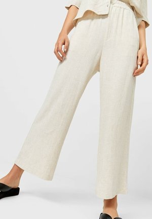 Woman wearing loose, high-waisted cream linen pants with elastic waistband and wide legs, paired with black slide sandals.