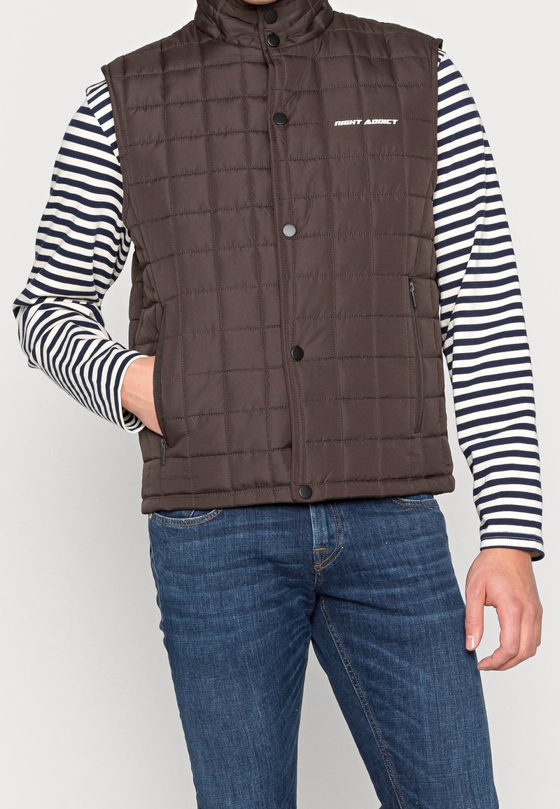 Man med en brun vadderad väst med tryckknappar över en långärmad svart och vit randig tröja och blå jeans, en hand i fickan.