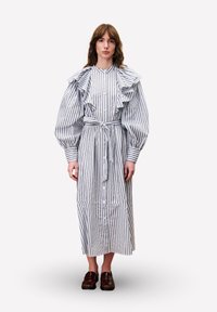 Eine Frau trägt ein langes, weißes Kleid mit vertikalen blauen Streifen, Rüschenschultern, Ballonärmeln, einem gebundenen Taillengürtel und dunkelbraunen Mokassins.