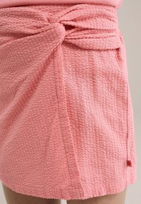 Rozā teksturēta apvalka svārki ar mezglu jostas vietā, kas rotāta ar mazu sarkanu birku. Audumam ir paaugstināta rakstu struktūra, kas piešķir papildu dimensiju.