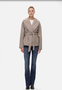 Béžový wrap kabát s širokým límcem, pásem v pase a dlouhými rukávy, nosený přes světlý top a kombinovaný s rozšířenými džíny a černými botami.