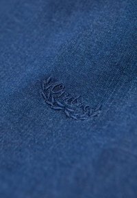 Tessuto blu scuro con una consistenza morbida, caratterizzato da un logo ricamato in un filo a contrasto. Il design presenta una leggera lucentezza e un motivo uniforme.