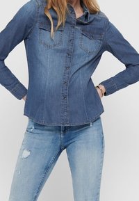 Kvinna som bär en figurnära denimskjorta med lång ärm och fickor fram, tillsammans med ljusa jeans med revor, stående med händerna på höfterna.