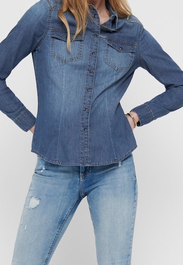 Kvinna som bär en figurnära denimskjorta med lång ärm och fickor fram, tillsammans med ljusa jeans med revor, stående med händerna på höfterna.