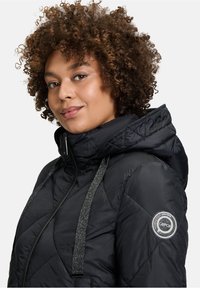 Sort quiltet jakke med hætte, glitrende stropper og et cirkulært logo. Materialet er glat med en let glans og mønstret syning.