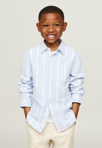 Camicia button-up a righe blu chiaro con taschino, realizzata in tessuto morbido. Maniche lunghe arrotolate, indossata sopra una camicia bianca e pantaloni beige.