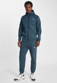 Tute con cappuccio in blu scuro, composto da una giacca con zip e tasche a marsupio e pantaloni affusolati coordinati; realizzato in materiale testurizzato.