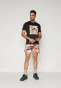 Homme en t-shirt graphique noir, shorts à motifs, chaussettes Nike grises et baskets noires tenant une bouteille d'eau noire, se tenant devant un fond uni.