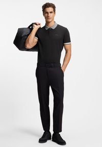 Czarna koszulka polo z wzorzystym kołnierzykiem, czarne spodnie i czarne trampki, trzymający czarną torbę. Na koszulce i pasku widoczne logo.